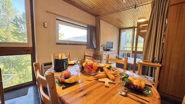 Una mesa de comedor de madera dispuesta con frutas, pan y una cafetera en una habitación con ventanas amplias y techo de madera.