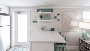A kitchen with a white island, teal chairs, a teal lamp, and a wall with decorative items including a sign that says 'to the beach I must go.'