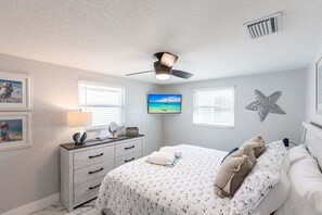 Une chambre à coucher comprenant un lit, une commode, un ventilateur de plafond, une télévision, une décoration en forme d’étoile de mer et des tableaux encadrés.