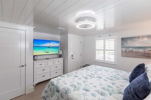 Une chambre à coucher avec un lit, une télévision, une commode, une fenêtre avec des stores et un tableau au mur.