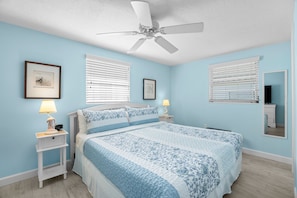Un dormitorio con cama, dos mesitas de noche con lámparas, ventilador de techo y dos ventanas con persianas.