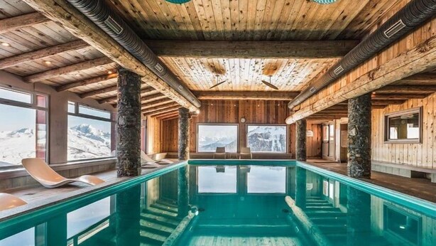 Uma grande piscina coberta com vigas de madeira e pilares de pedra, cercada por janelas que oferecem vista para as montanhas.