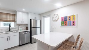 A modern kitchen with white cabinetry, stainless steel appliances, a white island, and a wicker chair. - Csa Blanca - Villa B18 (Sarasota)