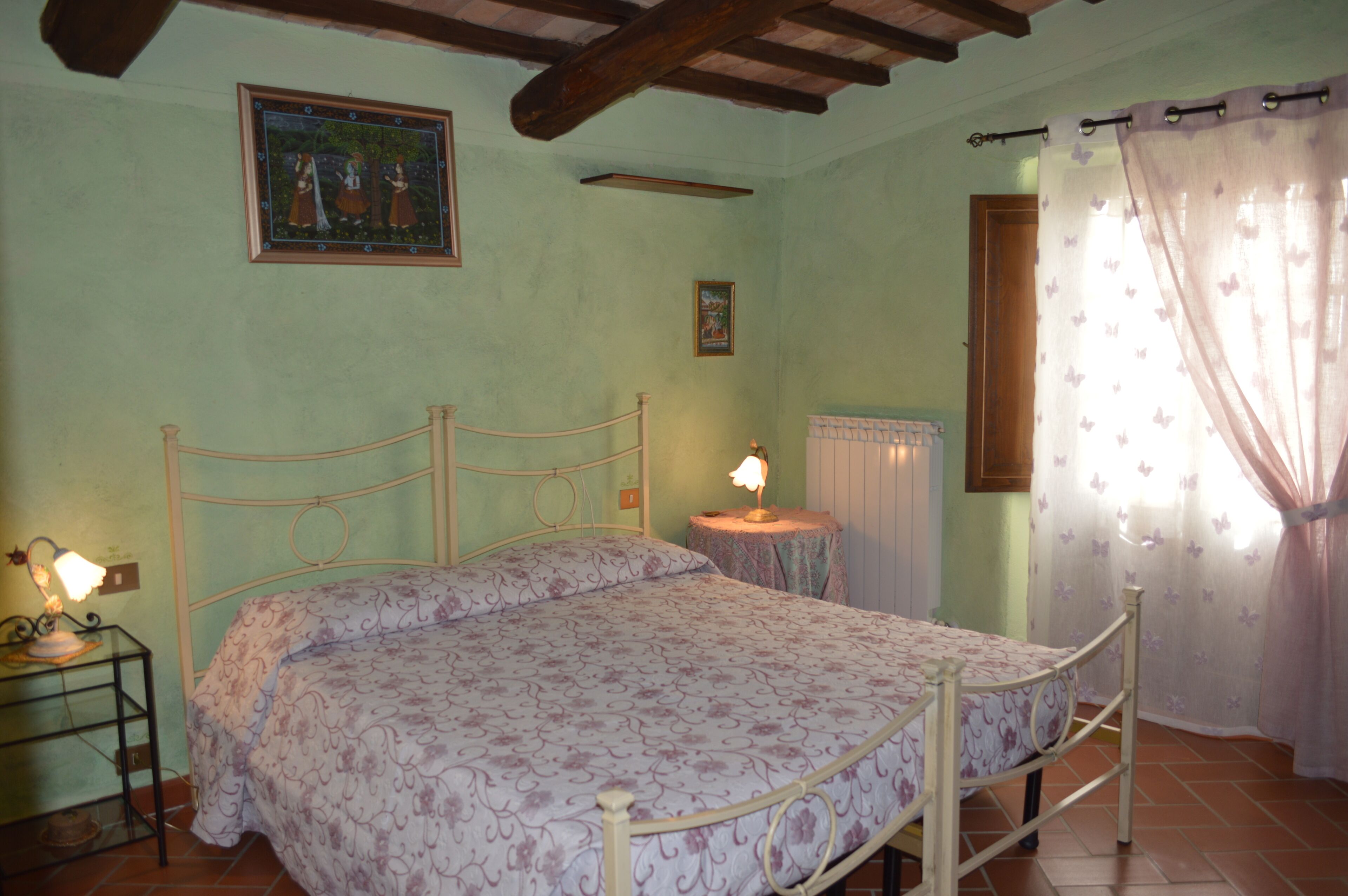 A bedroom with a bed, a bedside table with a lamp, a wall-mounted picture, and a window with curtains.