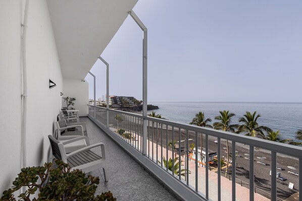 A balcony with white chairs and potted plants, overlooking a beach with palm trees and a clear sky.
