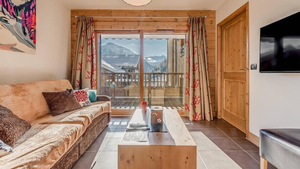 Ein gemütliches Wohnzimmer mit einer Couch, einem Couchtisch und einem Fernseher. Eine Holztür und ein Balkon mit Vorhängen sind zu sehen.