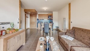 Eine moderne Küche mit Holzausstattung, ein Essbereich mit Tisch für vier Personen und ein Wohnzimmer mit gemustertem Sofa.