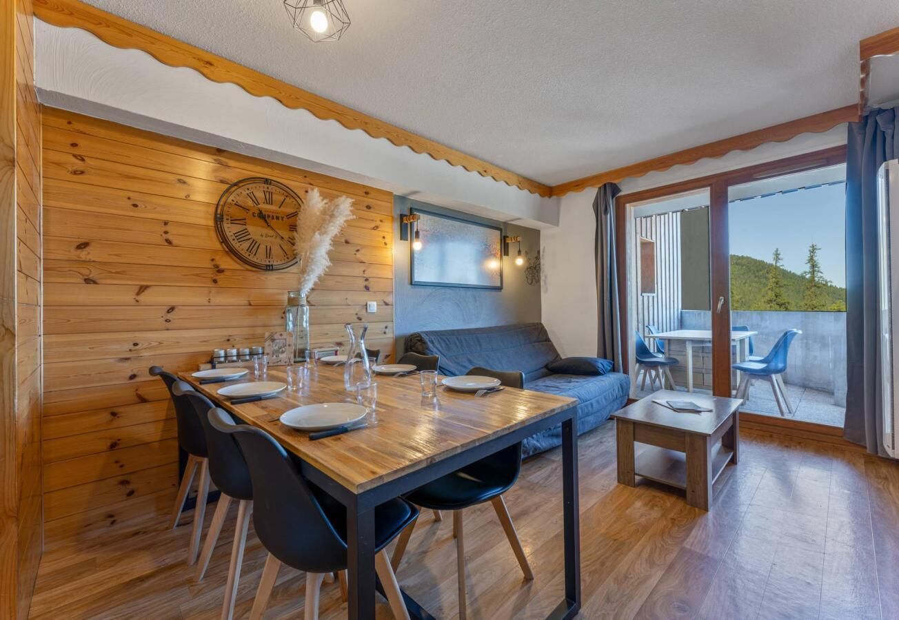 Ein gemütliches Blockhauszimmer mit einem Holzespeisetisch, gedeckt für vier Personen, einer Couch und einem kleinen Tisch mit Stühlen. Eine Uhr ist an der Wand befestigt, und es gibt ein Fenster mit Vorhängen.