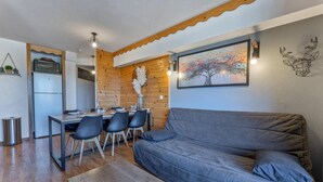 A cozy living area with a grey sofa, wooden dining table, and chairs. A small kitchen with a refrigerator and a microwave is visible.