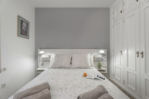 A neatly arranged bedroom with a bed, bedside lamps, a nightstand, a framed picture, and a wardrobe.