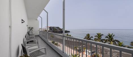 A balcony with white chairs and potted plants, overlooking a beach with palm trees and a clear sky.