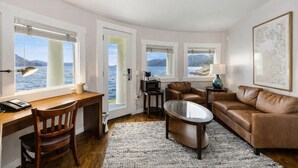 A modern living room with a wooden desk, leather sofa, and a glass coffee table. Large windows offer a view of the sea and mountains.