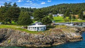 A coastal property with a circular building, rocky shoreline, clear water, and a forested hill in the background.