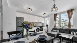 Una cocina moderna con armarios blancos, un comedor con una mesa para cuatro personas y una sala de estar con sofá y vista a un edificio por la ventana.