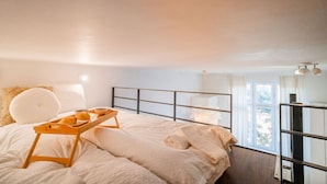 Une chambre à coucher avec un lit, une table de chevet sur laquelle se trouve une assiette de pain, une fenêtre avec des rideaux et une vue sur l’extérieur.