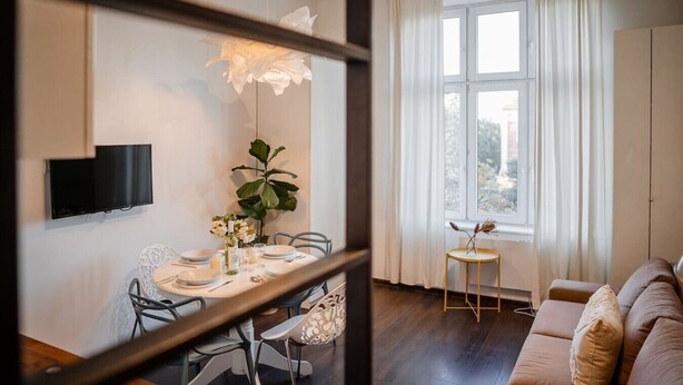Un coin repas avec une table dressée pour deux personnes, une plante en pot et une petite table ronde sur laquelle se trouve un vase. On aperçoit un canapé et une fenêtre avec des rideaux au fond.