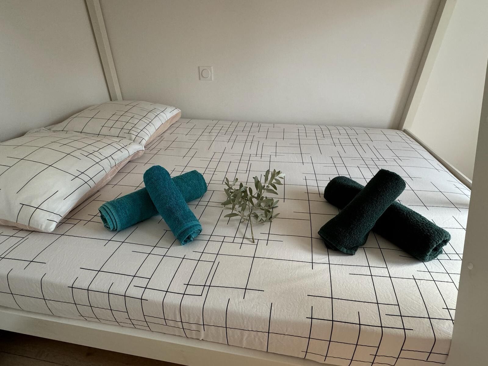 A bed with a grid-patterned bedspread, two rolled towels, a decorative plant, and a pillow with a geometric design.