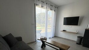 Un salon comprenant un canapé, un banc en bois, un téléviseur fixé au mur et une porte-fenêtre coulissante donnant sur un espace extérieur.