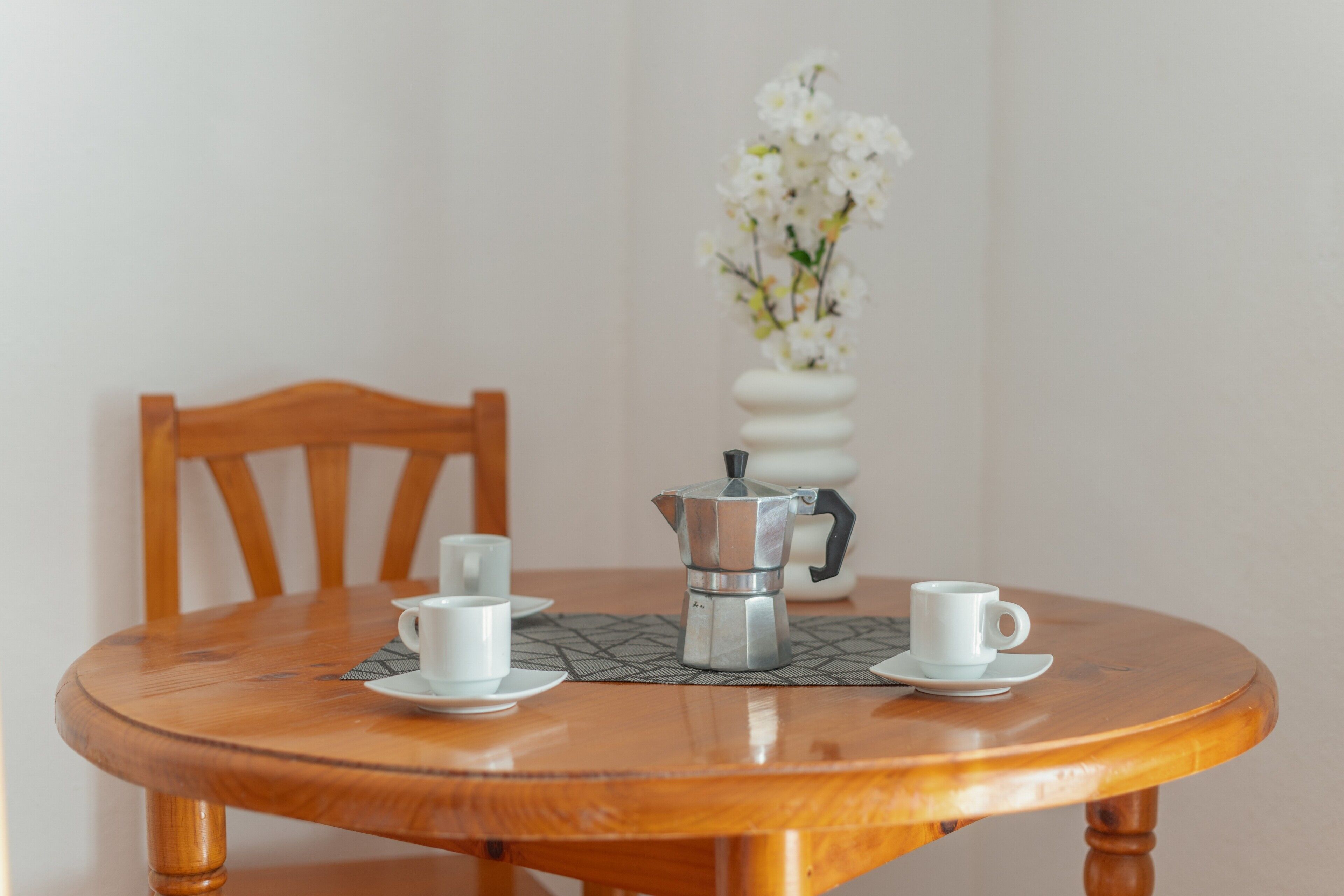 Un tavolo rotondo in legno con una caffettiera e due tazze, una sedia e un vaso con fiori bianchi.