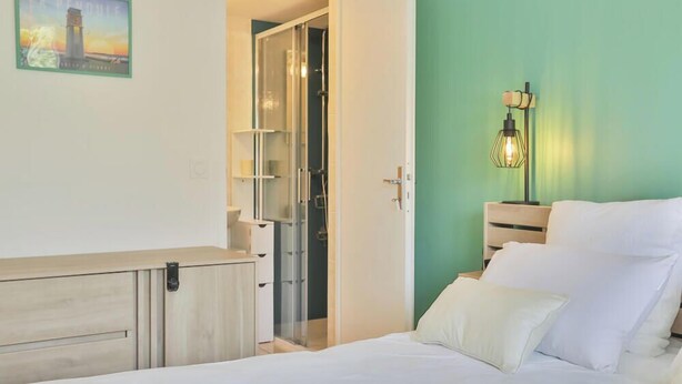 A bedroom with a bed, a bedside table with a lamp, a wardrobe, and a bathroom visible through an open door. - Grande Maison avec Jardin, Parking, proche de la Plage (Les Sables-d'Olonne)