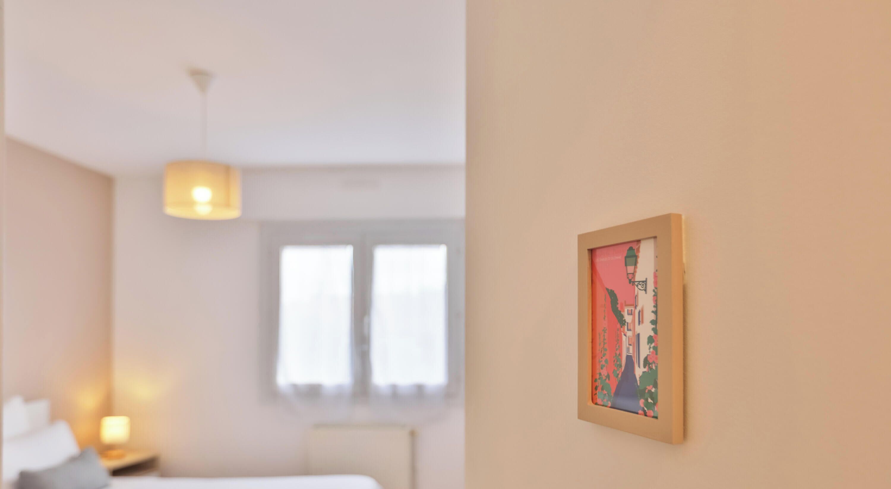 Una habitación de hotel con cama, mesita de noche, lámpara, ventana y un cuadro en la pared.