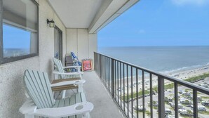 A balcony with two Adirondack chairs, a small table, and a window overlooking a beach with parked vehicles.