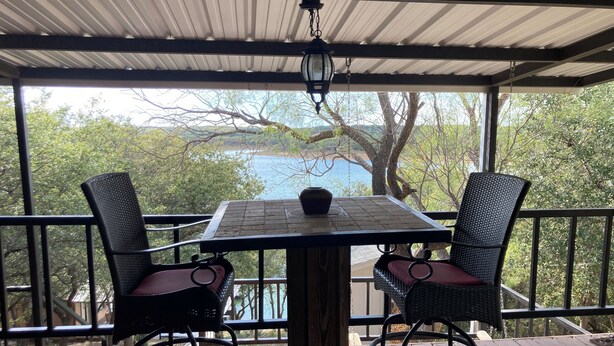 Un área de estar al aire libre con techo, con una mesa y sillas, con vistas a un paisaje de árboles y una masa de agua.