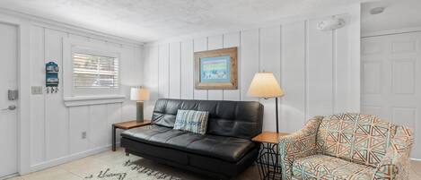 Un salon avec un canapé en cuir noir, un fauteuil à motifs, un tapis, un tableau encadré au mur et une lampe sur une table d’appoint.