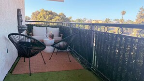 En balkon med en hvid sofa, to sorte stole, et lille bord med en flaske og et glas, samt en rød måtte.