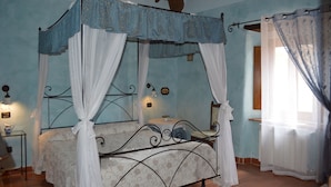 A four-poster canopy bed with white drapes, a small table with a lamp, and a chair in a room with blue walls and wooden beams.