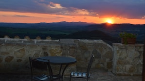 A stone wall with a table and chairs, overlooking a sunset over a hilly landscape.
