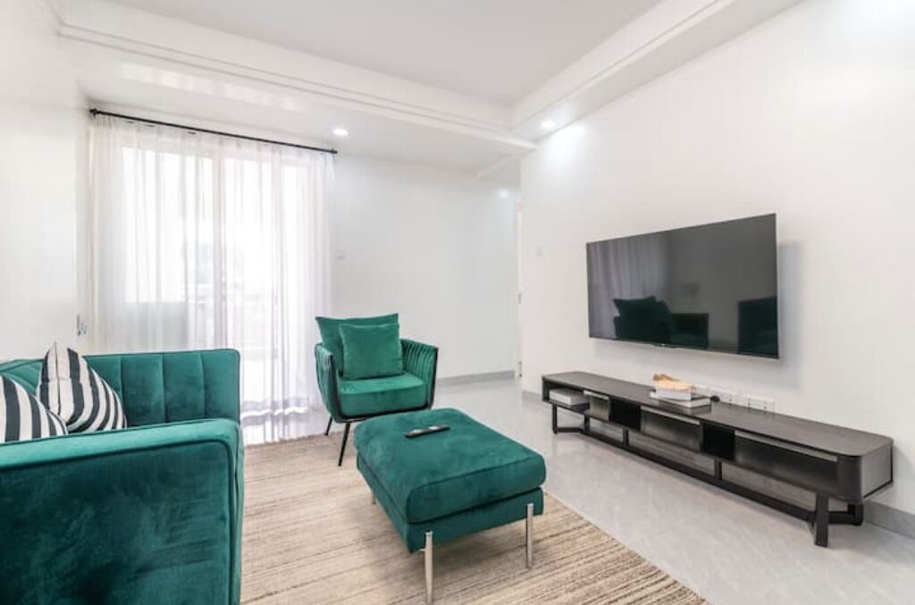 A modern living room with a green sofa, matching ottoman, and a green armchair. A flat-screen TV is mounted on the wall, and a dark wooden console table is against the wall.