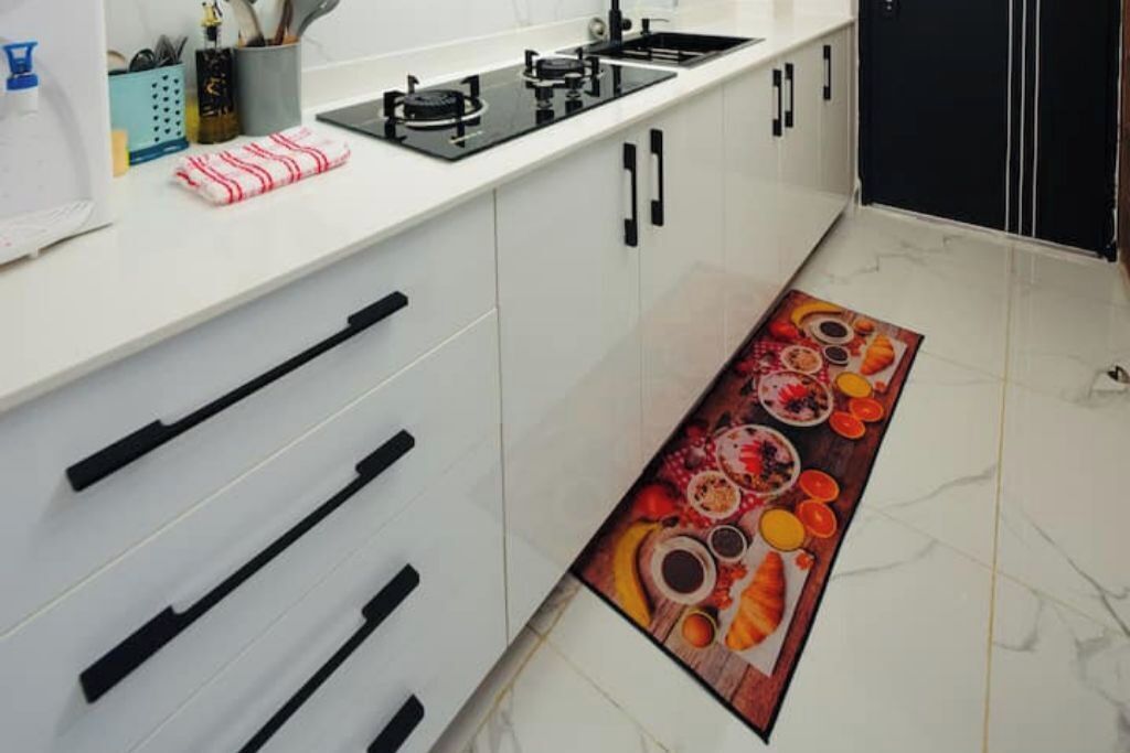 Una cocina con armarios blancos, pomos negros, una estufa negra y una alfombra colorida que muestra alimentos.