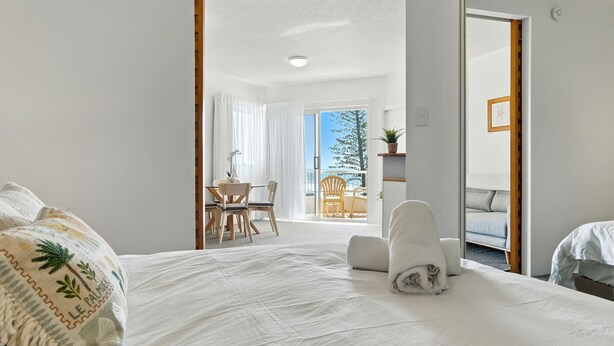 Een slaapkamer met een groot bed, een kleine tafel met stoelen en een balkon met uitzicht op de lucht en bomen.
