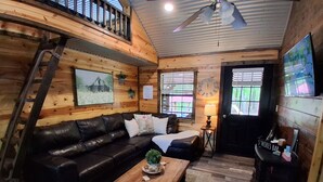 A cozy cabin interior with a wooden floor, a black leather sofa, a wooden coffee table, a ladder, a clock, and a bar area.