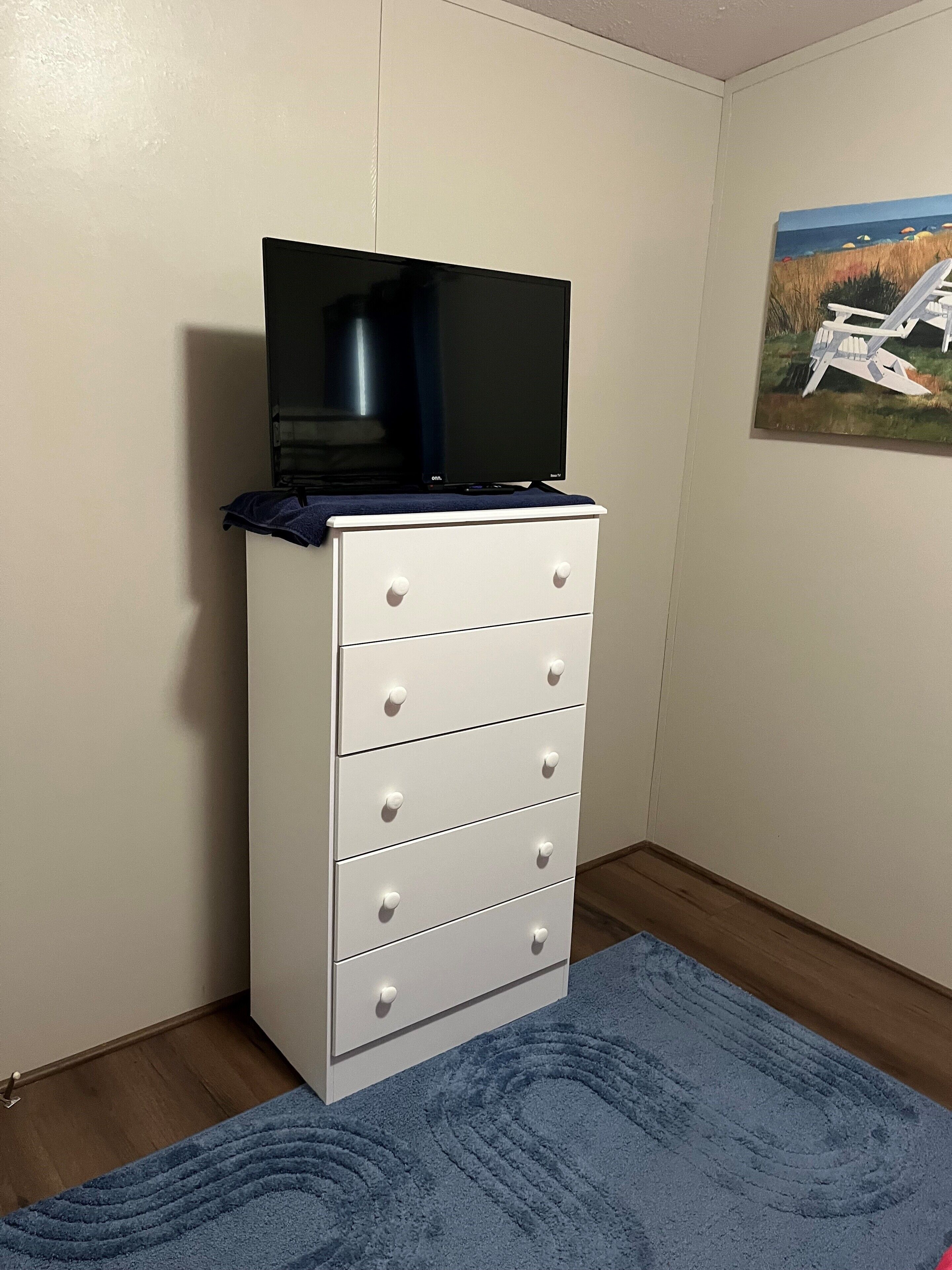 A white dresser with a flat-screen TV on top, a blue rug on the floor, and a painting of beach chairs on the wall.