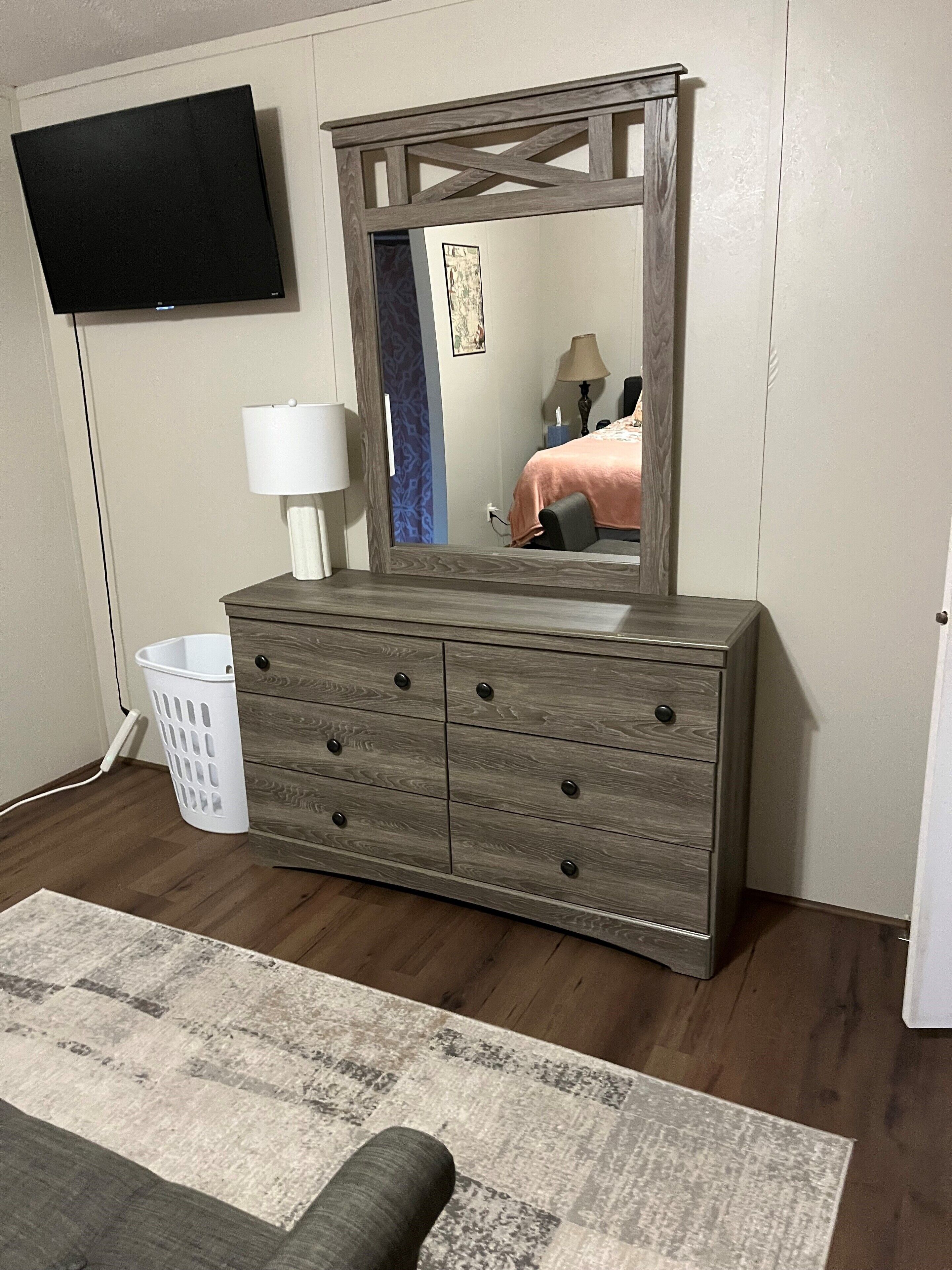 A bedroom with a wooden dresser, a television mounted on the wall, a white lamp, and a patterned rug.