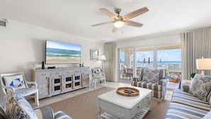 A living room with a television, white furniture, a ceiling fan, and a view of the ocean. - Sandpiper 108 (New Smyrna Beach)