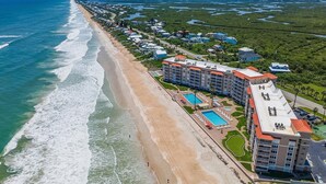 A coastal hotel with a long beachfront, multiple swimming pools, and a large building with a red roof.