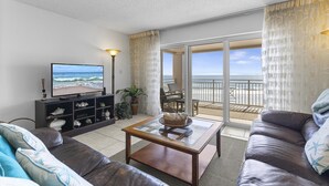 A living room with a television, a sofa, a coffee table, and a balcony with a view of the beach.