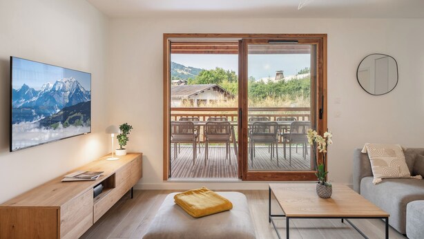 Ein modernes Wohnzimmer mit großem Fenster, Holzmöbeln, einem Flachbildfernseher und Blick auf eine Berglandschaft sowie einen Sitzbereich im Freien.