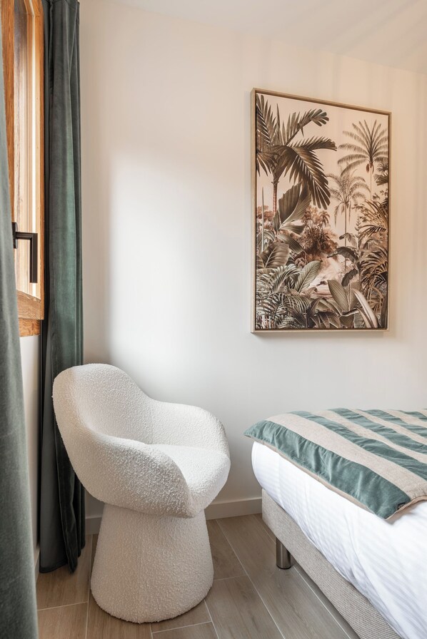A bedroom with a bed, a white chair, a tropical-themed painting, and a window with curtains.