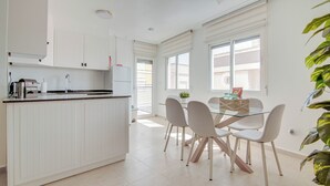 A modern kitchen with white cabinetry, a glass dining table, and a view of the city through large windows. - 50m from the beach | Calle San Enrique 3-2 (Los Alcázares)