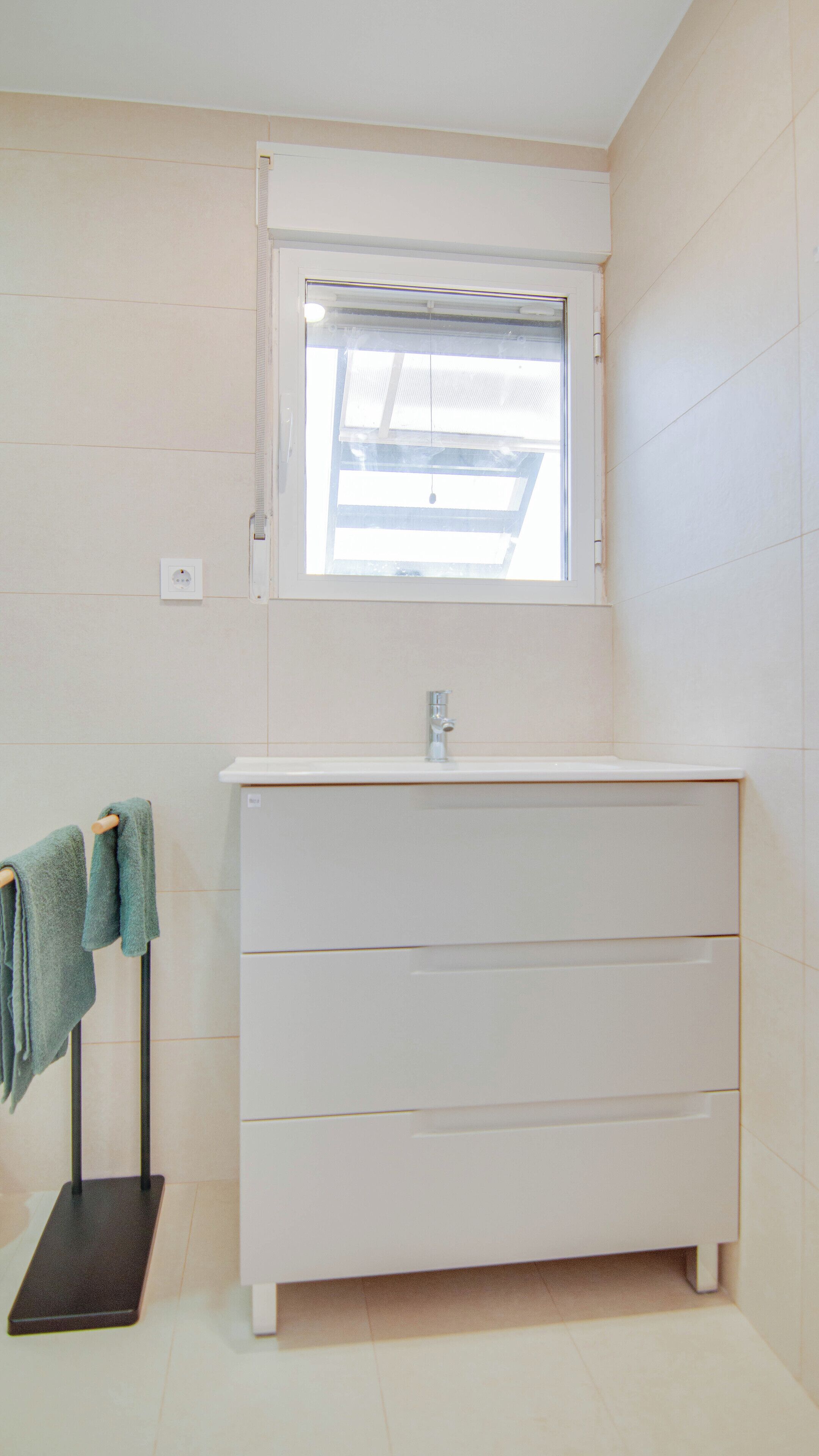Un baño con tocador blanco, una ventana sobre el lavabo y un perchero con dos toallas.