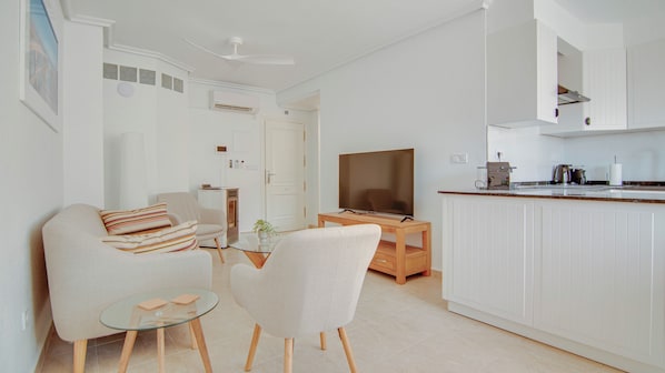 A modern, well-lit living room with a sofa, two armchairs, a glass coffee table, and a TV. A kitchen area with white cabinets and a countertop is visible.
