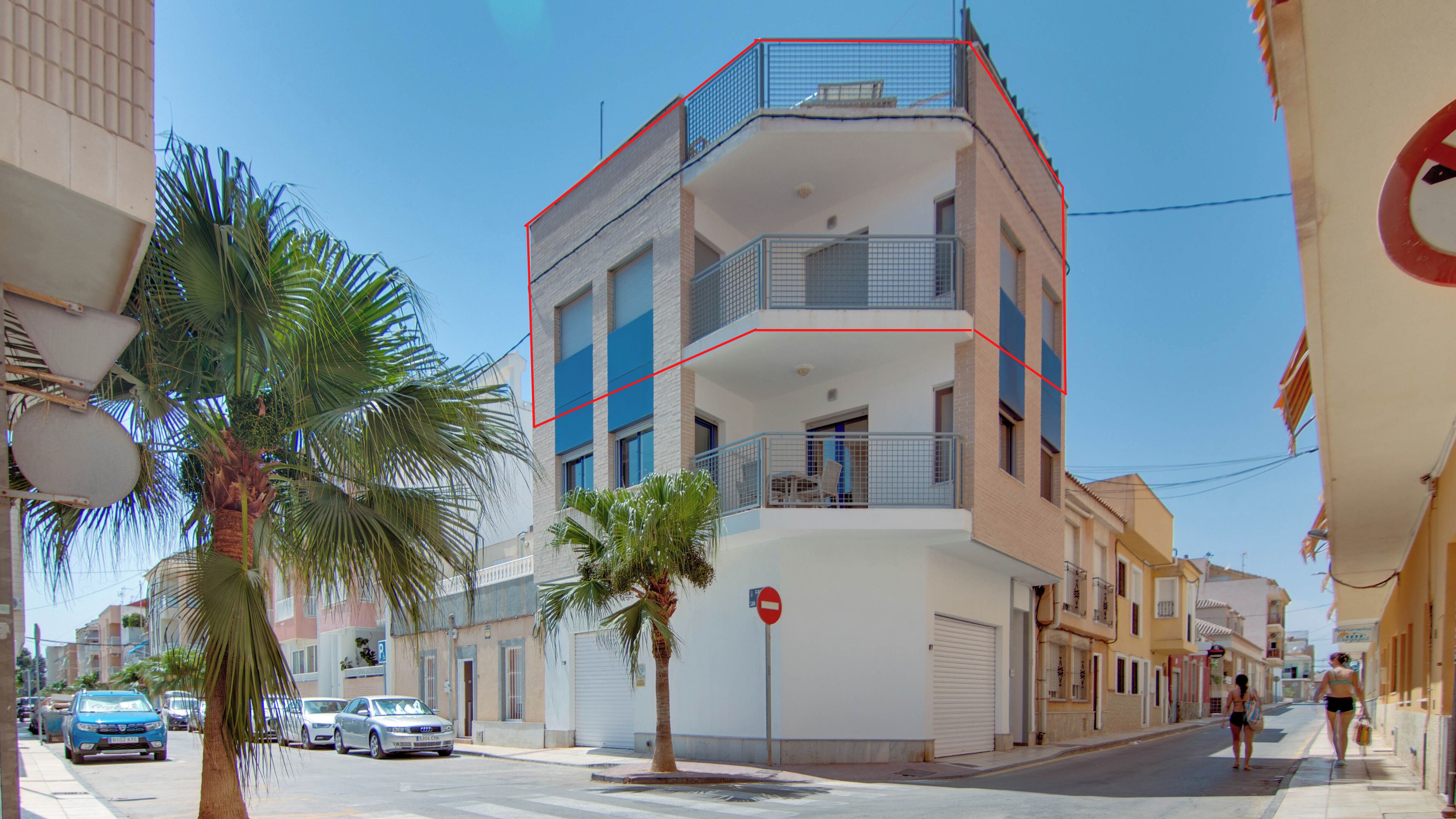 A multi-story building with balconies, a no entry sign, and parked cars on a sunny street.