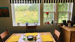 A dining area with a wooden table, yellow placemats, and chairs. There are windows with views of greenery outside. - The Bumbletown Hill House (Allouez)
