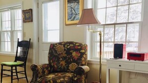 A room with a floral armchair, a wooden chair, a desk with a lamp, and a window with a view of trees. - The Bumbletown Hill House (Allouez)