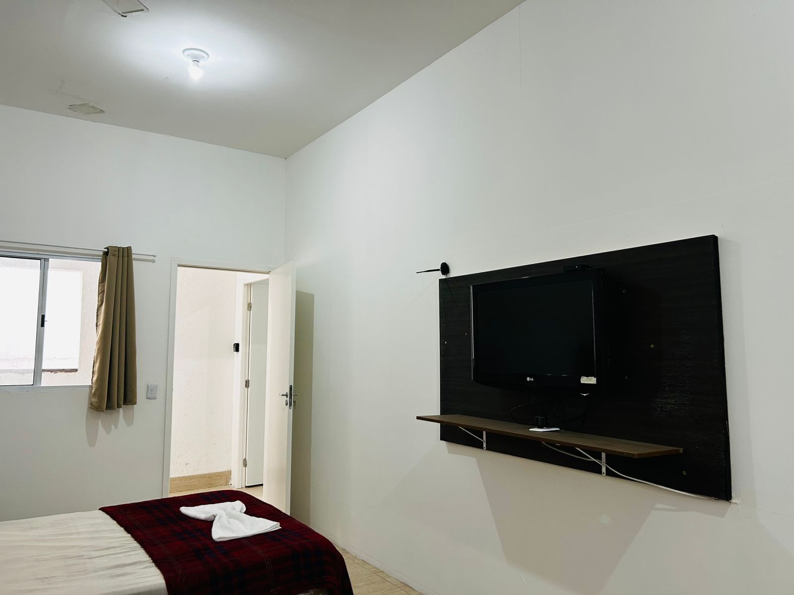 A bedroom with a bed, a TV mounted on the wall, a window with curtains, and a door.