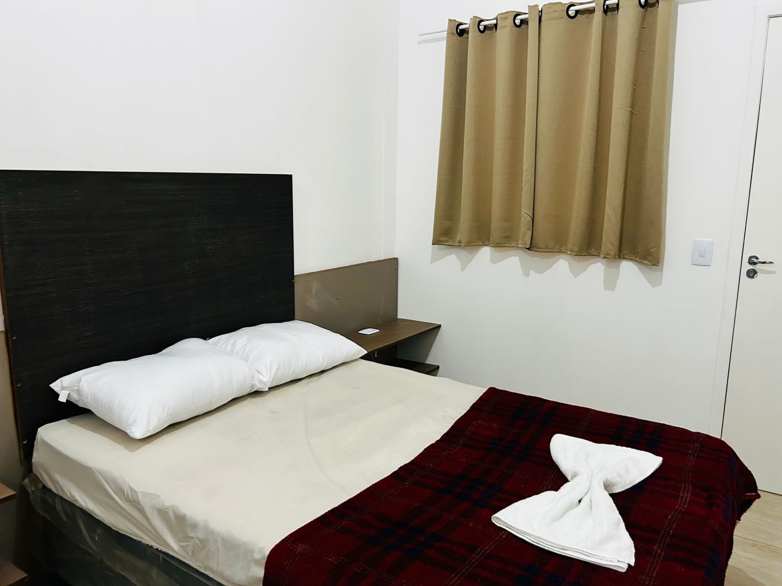 Ein Einzelbett mit weißer Bettwäsche, einem dunklen Kopfteil, einer roten Karo-Decke und einem weißen Handtuch.